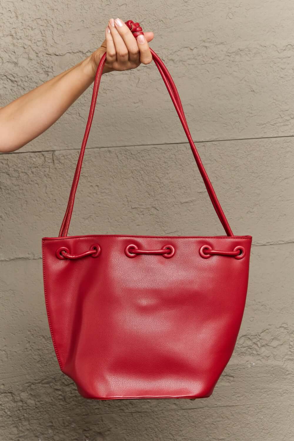 Red vegan leather bucket bag with drawstring closure held by a person against a textured wall.