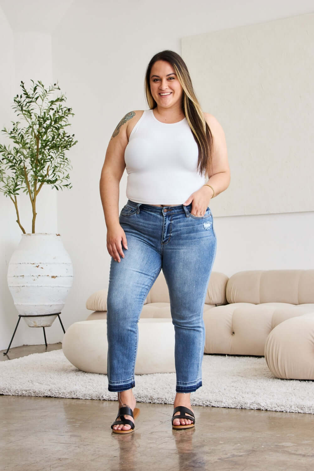 Woman modeling Judy Blue release hem cropped bootcut jeans in a modern living room setting.