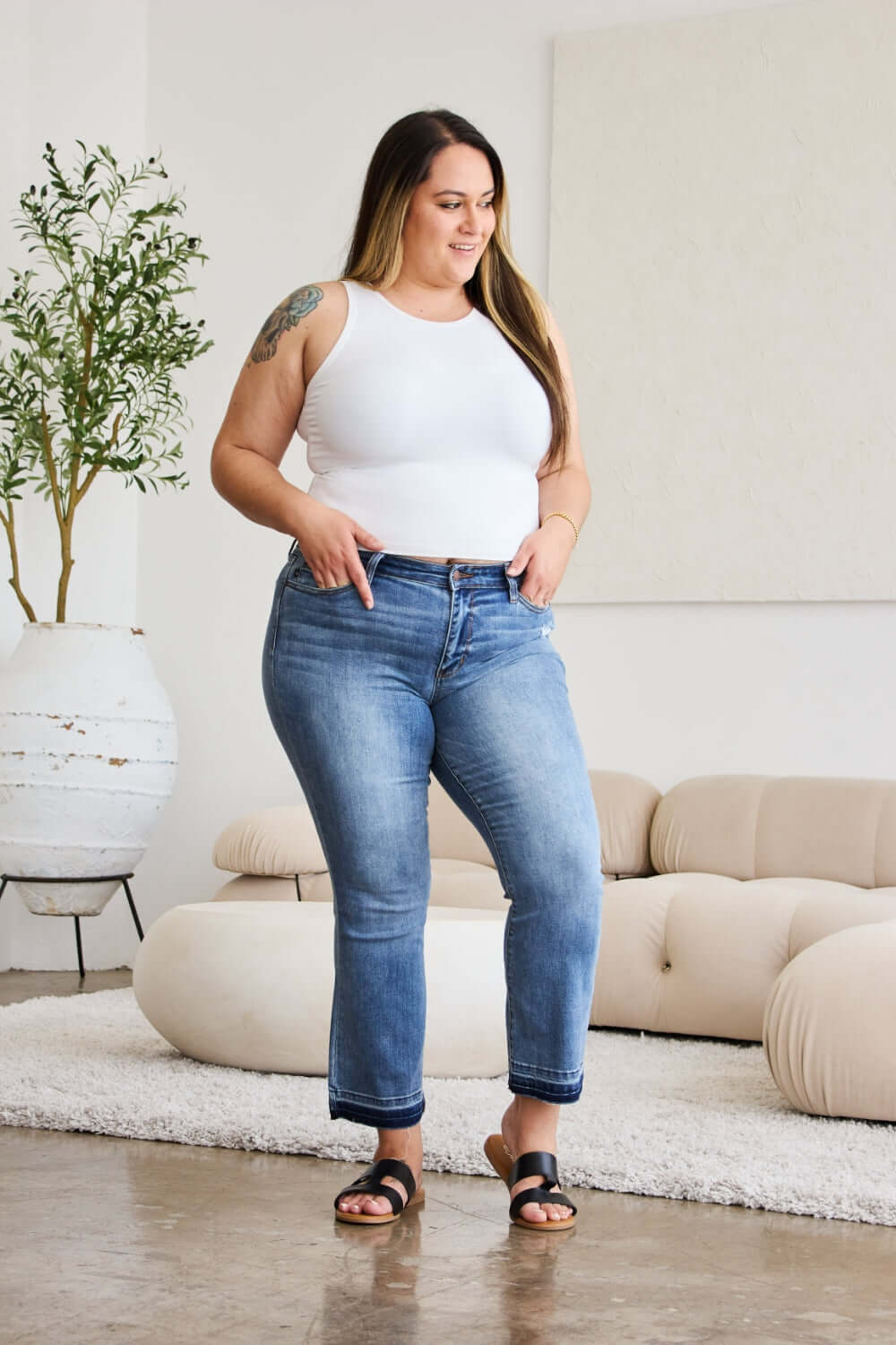 Woman wearing Judy Blue release hem cropped bootcut jeans in a modern living room.