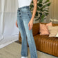 Woman wearing Judy Blue full size medium rise bootcut jeans with a casual white top and white sneakers in a modern living room setting.