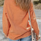 Woman wearing Bella Road THANKFUL Round Neck Long Sleeve Sweatshirt in orange, paired with blue jeans and a floral crossbody bag, standing outdoors.