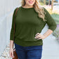 Plus-size model wearing a green round neck batwing sleeve top with jeans, accessorized with sunglasses and a brown shoulder bag.