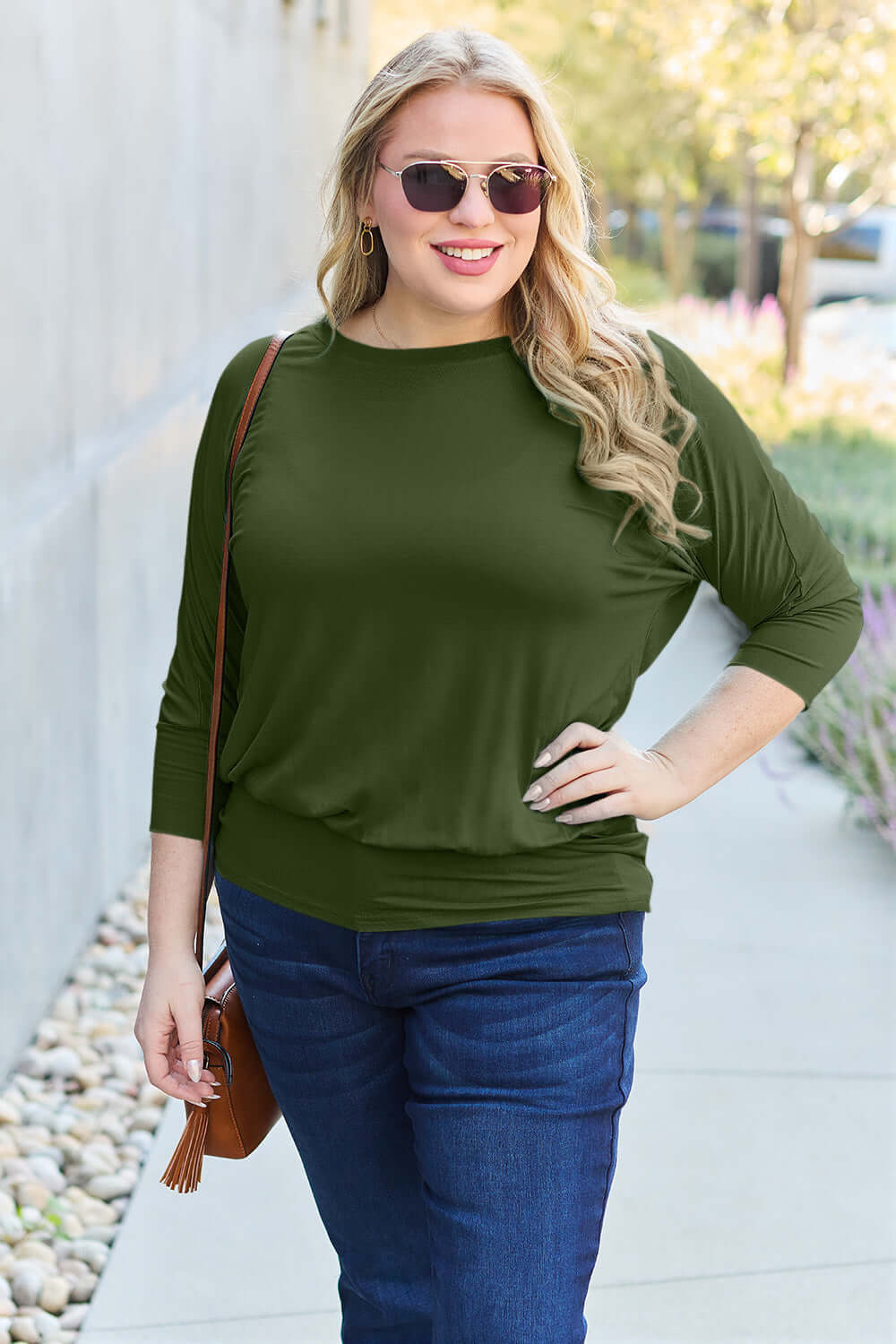 Plus-size model wearing a green round neck batwing sleeve top with jeans, accessorized with sunglasses and a brown shoulder bag.