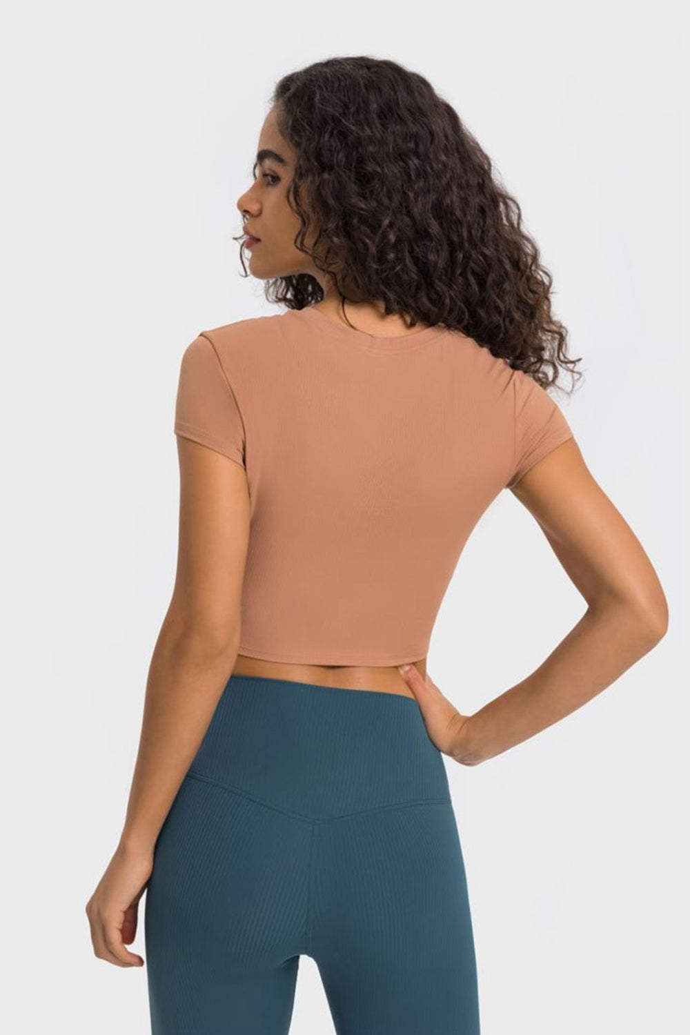 Back view of a model wearing a cropped short sleeve sports t-shirt in tan, paired with teal leggings for a stylish workout look.