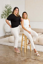 Two women showcase chic white jeans in a modern, comfy setting, one standing and the other seated on a wooden stool.