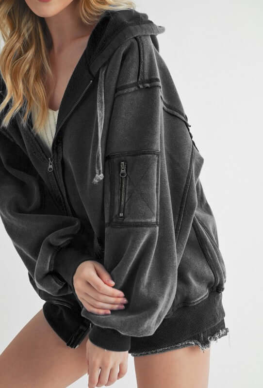 Trendy black zip-up drawstring hooded jacket with exposed seams and stylish pocket detail, perfect for cool weather.