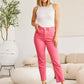 Woman wearing pink RFM jeans with high waist and raw hem, paired with white tank top and black heels, standing in modern living room.